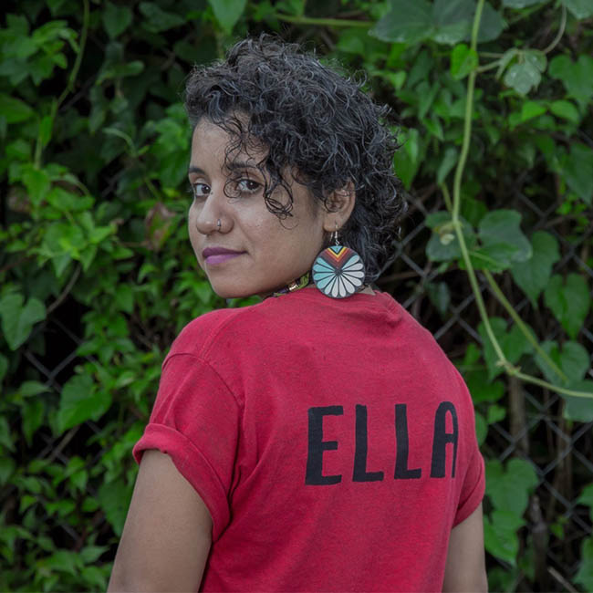 Yudith stands with their back towards the camera, face facing forward.. They are standing in front of green vines. They're wearing blue, yellow, and white circular earrings and a read shirt that says "ELLA" in black letters.
