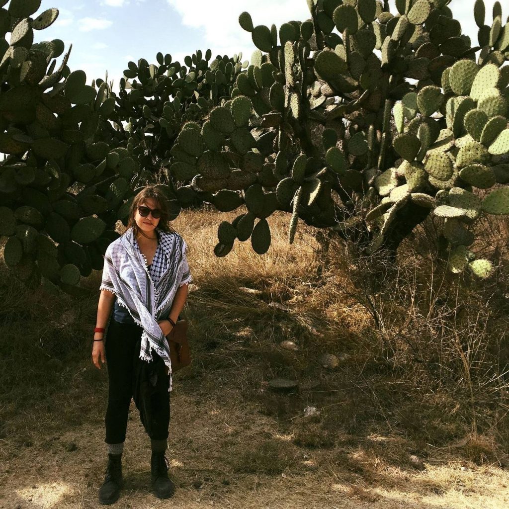 Sabina stands in a field in front of several big cacti plants. They're wearing black pants, sunglasses, and a white and black checkered scarf.